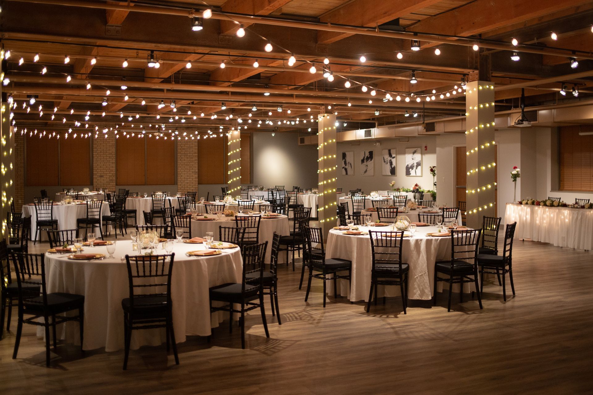 Elegant banquet hall with round tables, black chairs, and warm string lights hanging from the wooden ceiling.