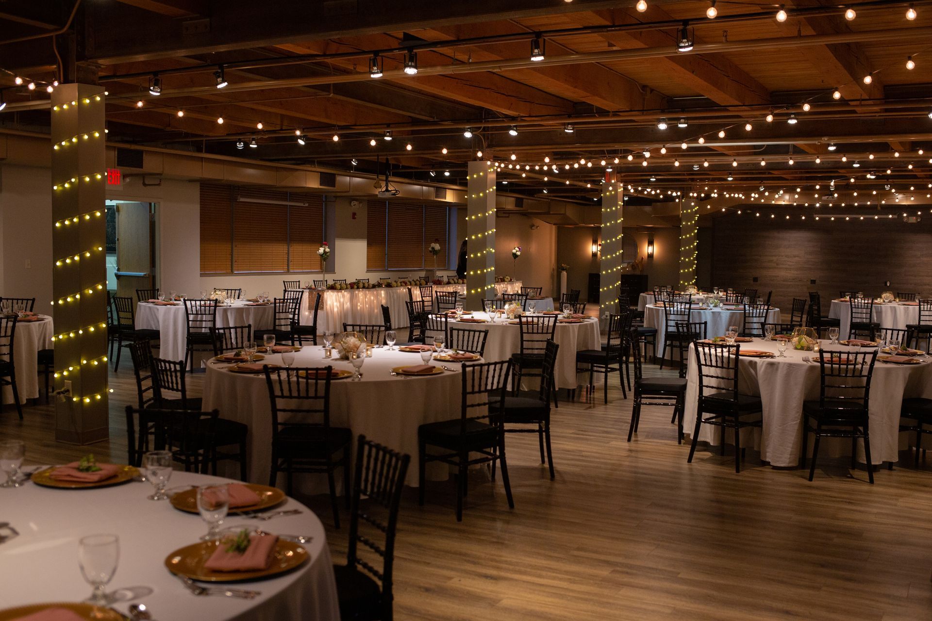 Empty banquet hall with round tables, black chairs, and warm string lights under exposed wooden beams