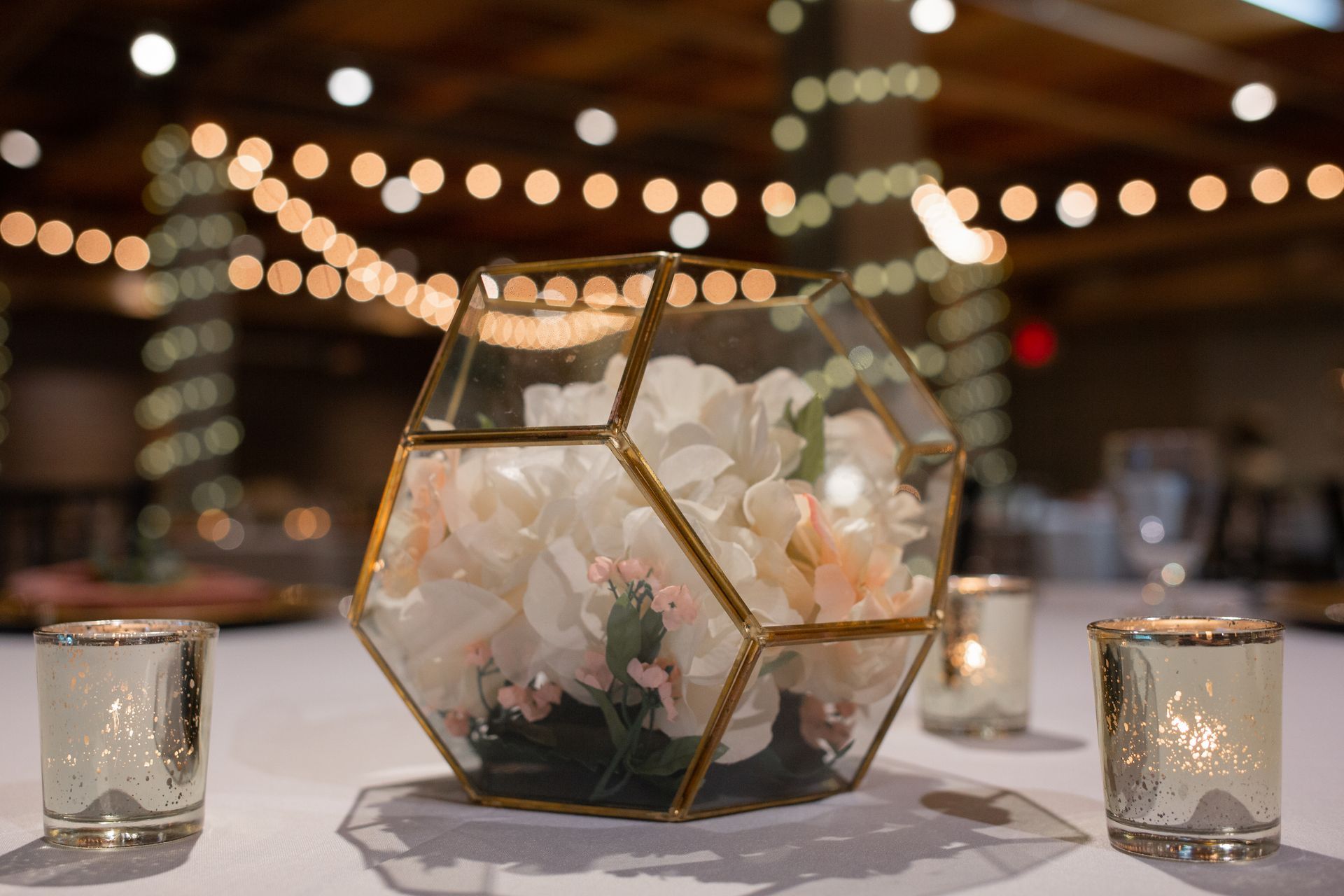 Gold geometric floral centerpiece with white flowers and candles on a decorated table, with warm lights in the background