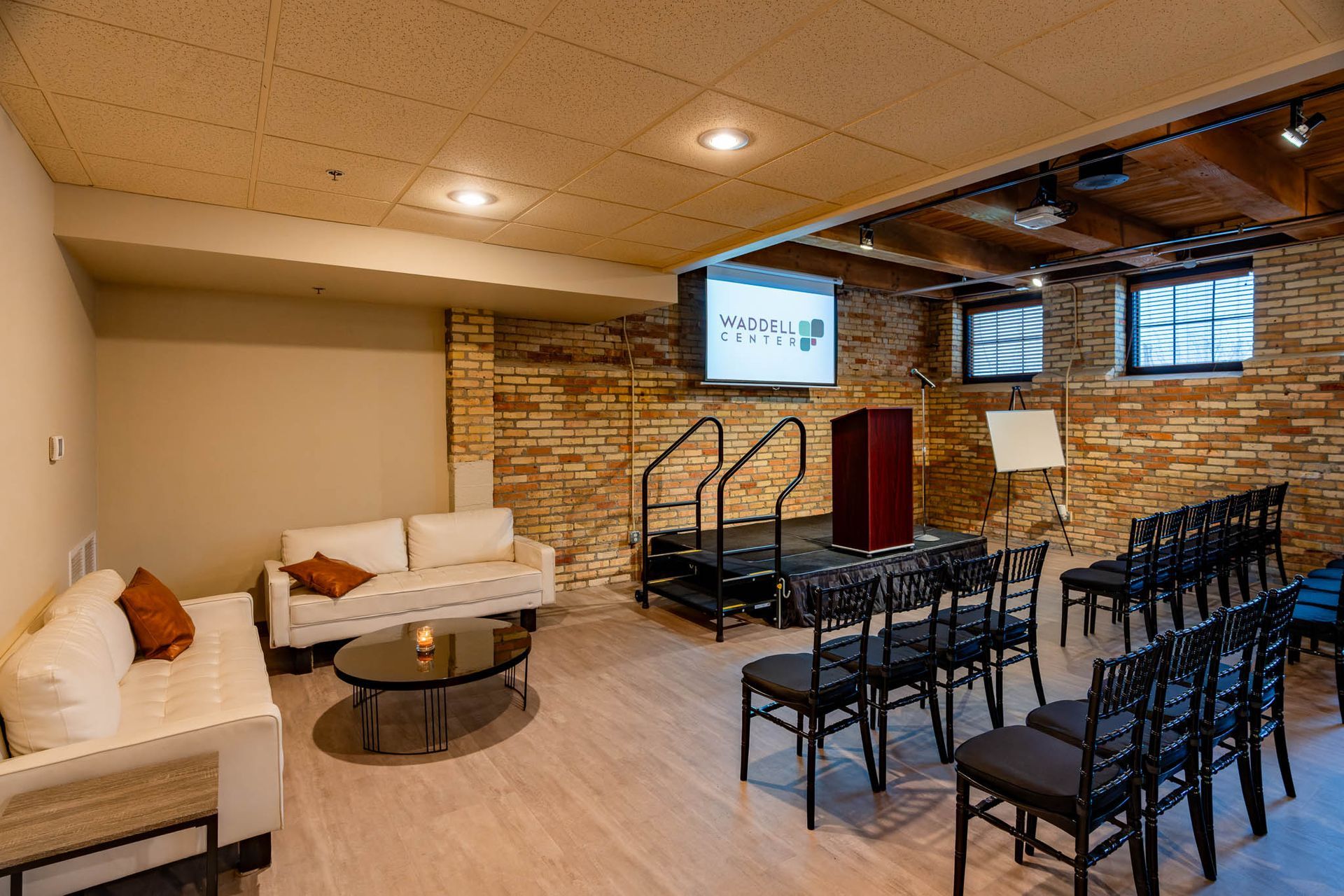 Lounge with white couches, black chairs, round table, and projector screen in a brick-walled room