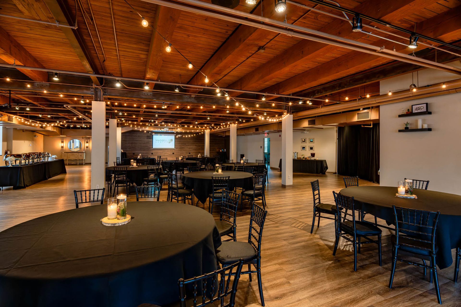 Empty restaurant dining room with round tables, black chairs, and warm wood ceiling lighting