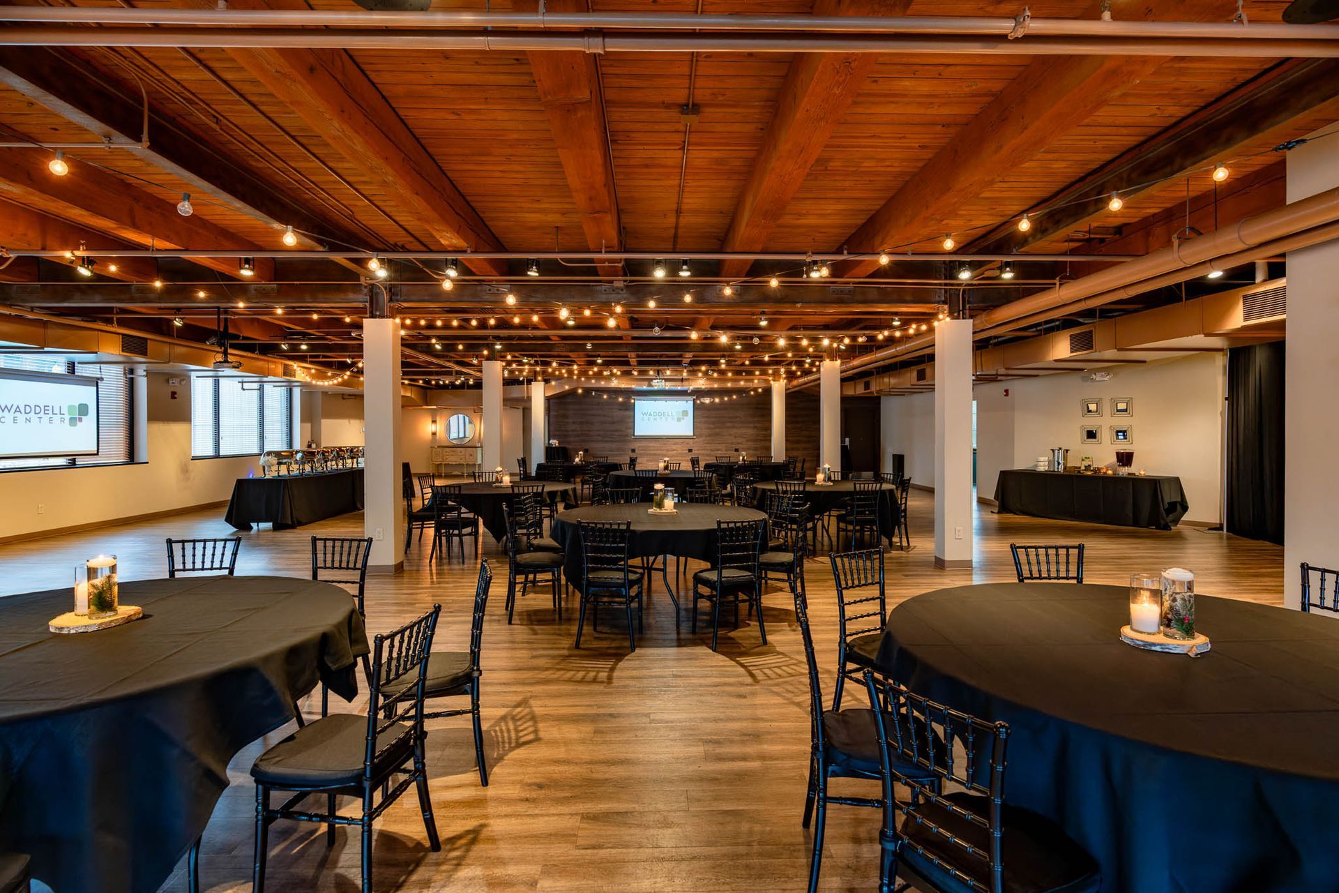 Spacious rustic event hall with round tables, black chairs, exposed wood beams, and warm lighting.