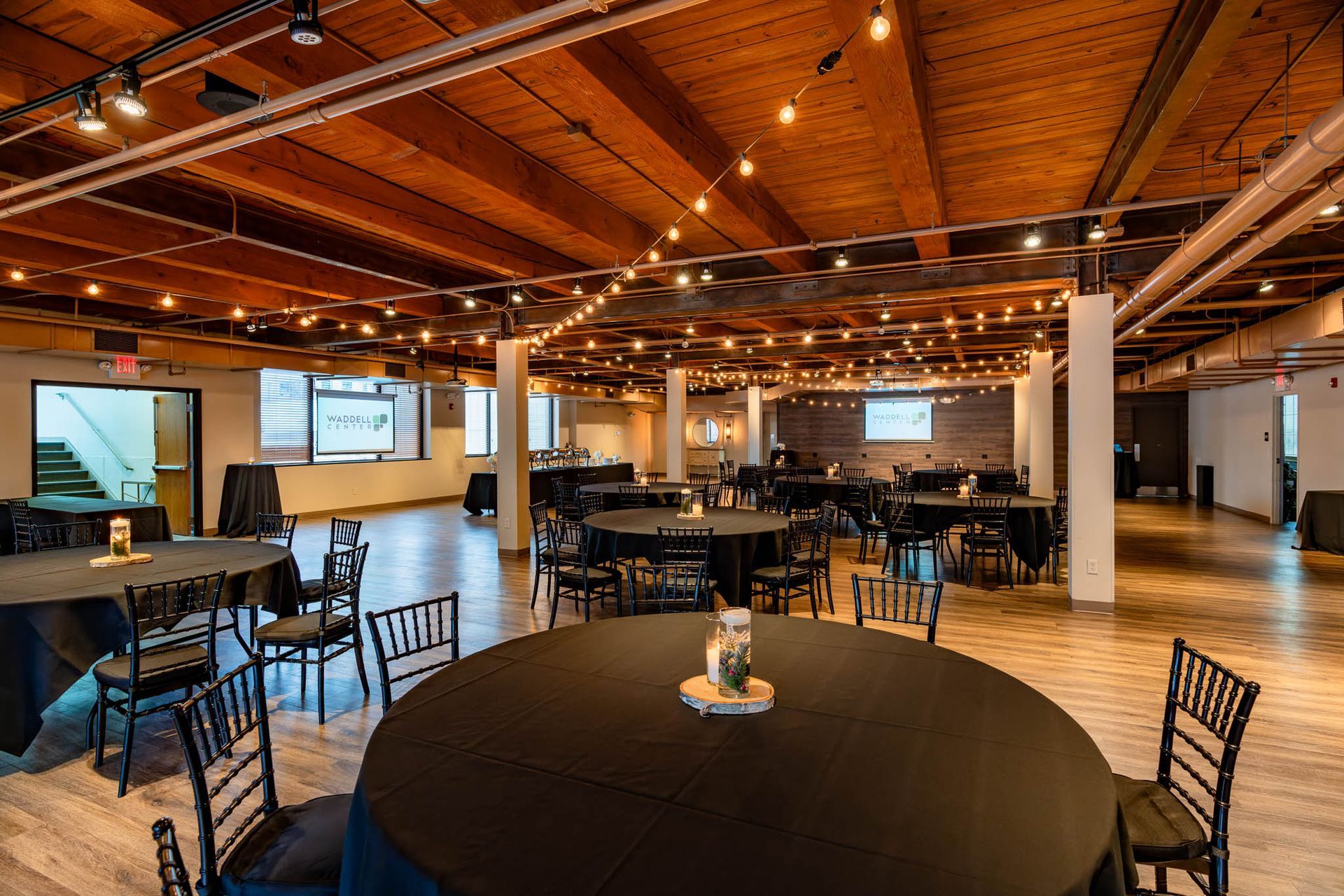 Spacious event hall with round tables, black chairs, wood beam ceiling, and warm string lights