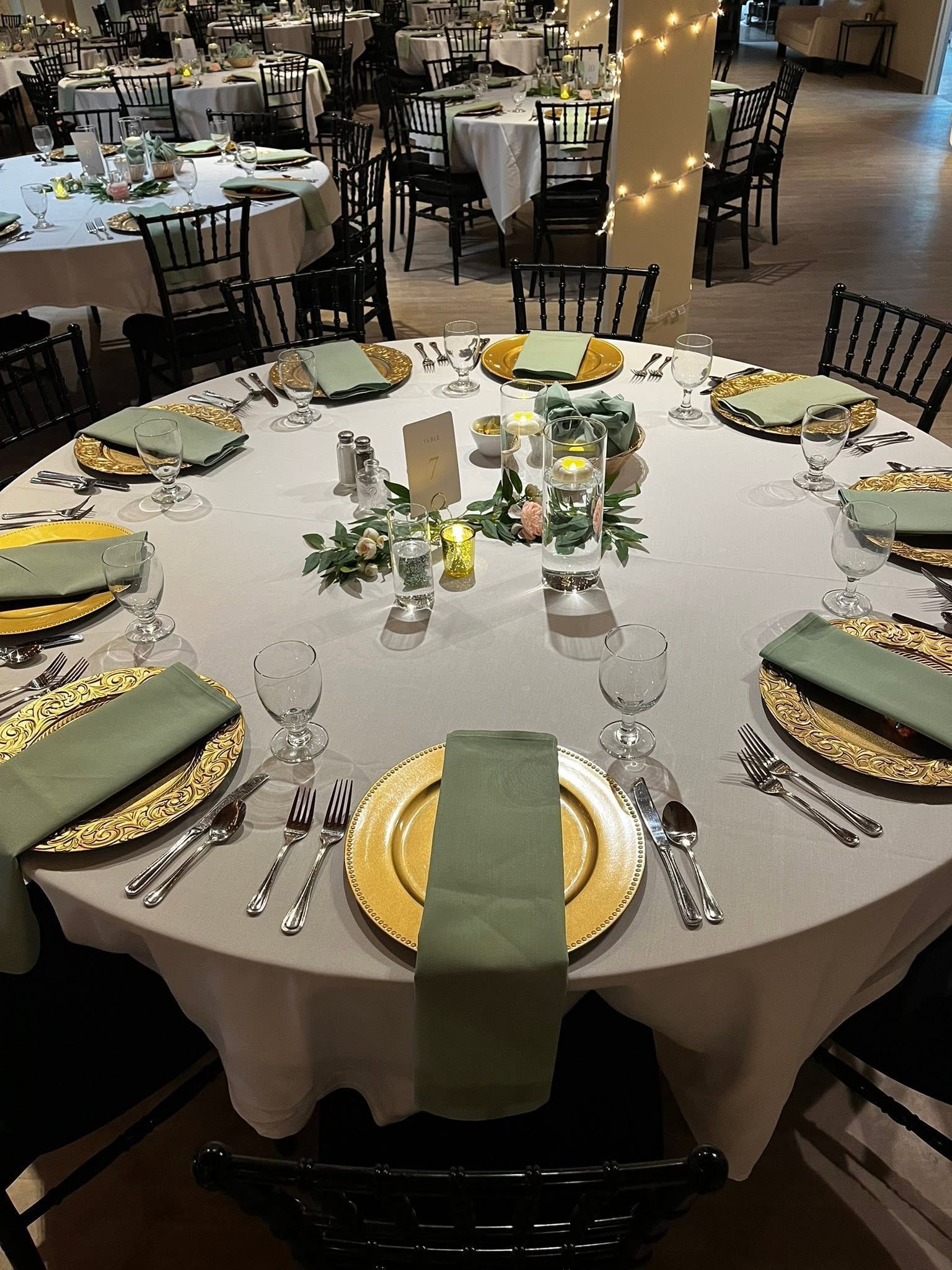 Round banquet table set with white linens, gold plates, green napkins, and candles in a decorated event hall