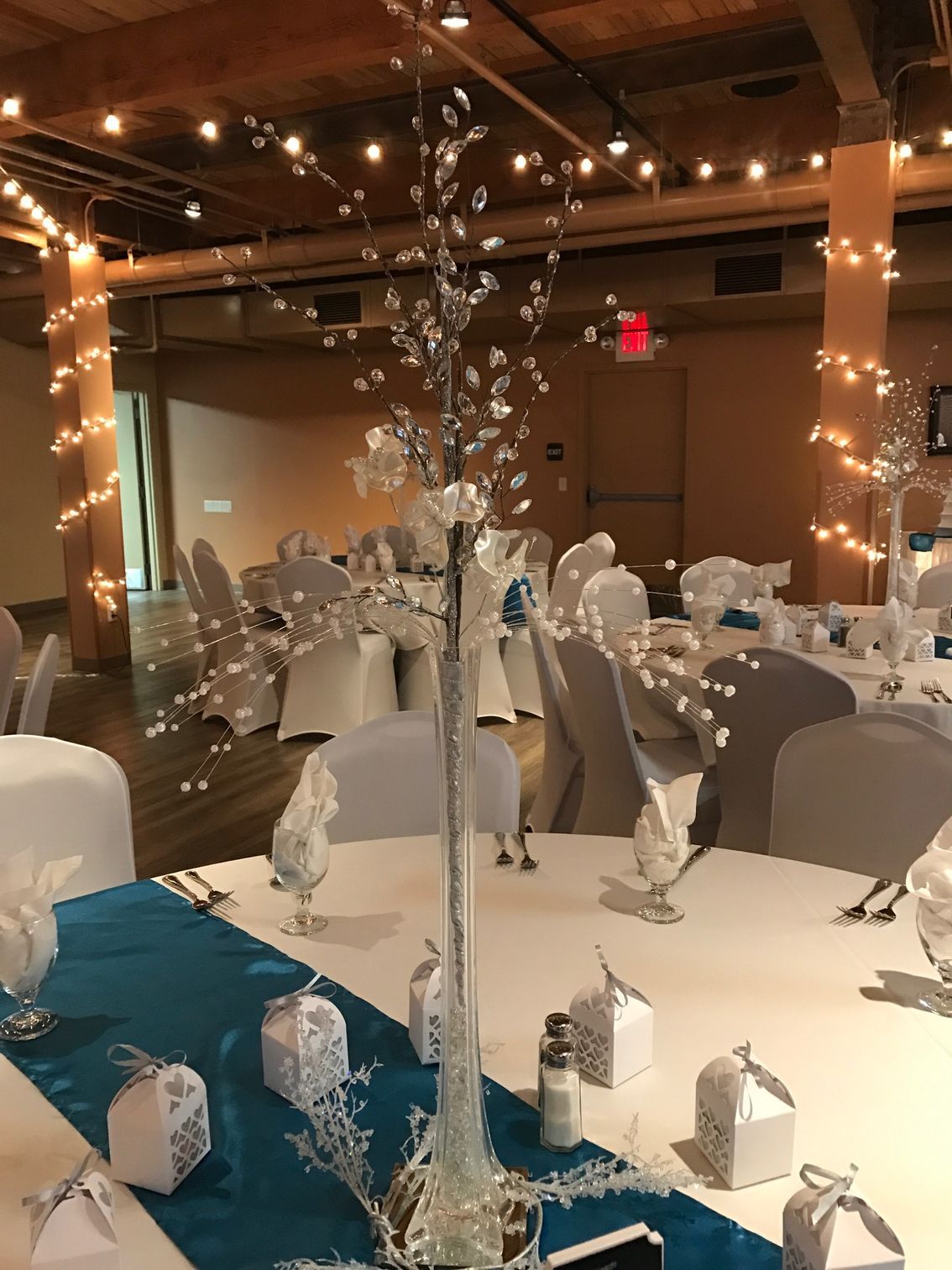 Decorated banquet hall with white-clothed tables, silver centerpieces, and string lights under wooden beams