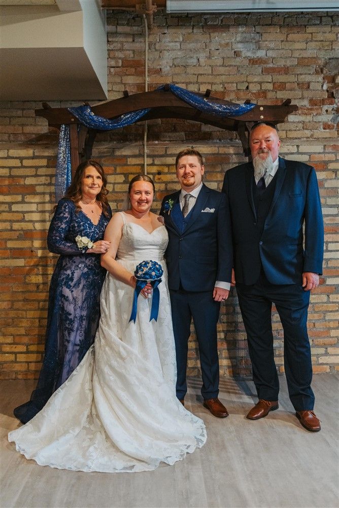 Wedding party posing indoors in front of a brick wall, with the bride in a white gown and blue bouquet.