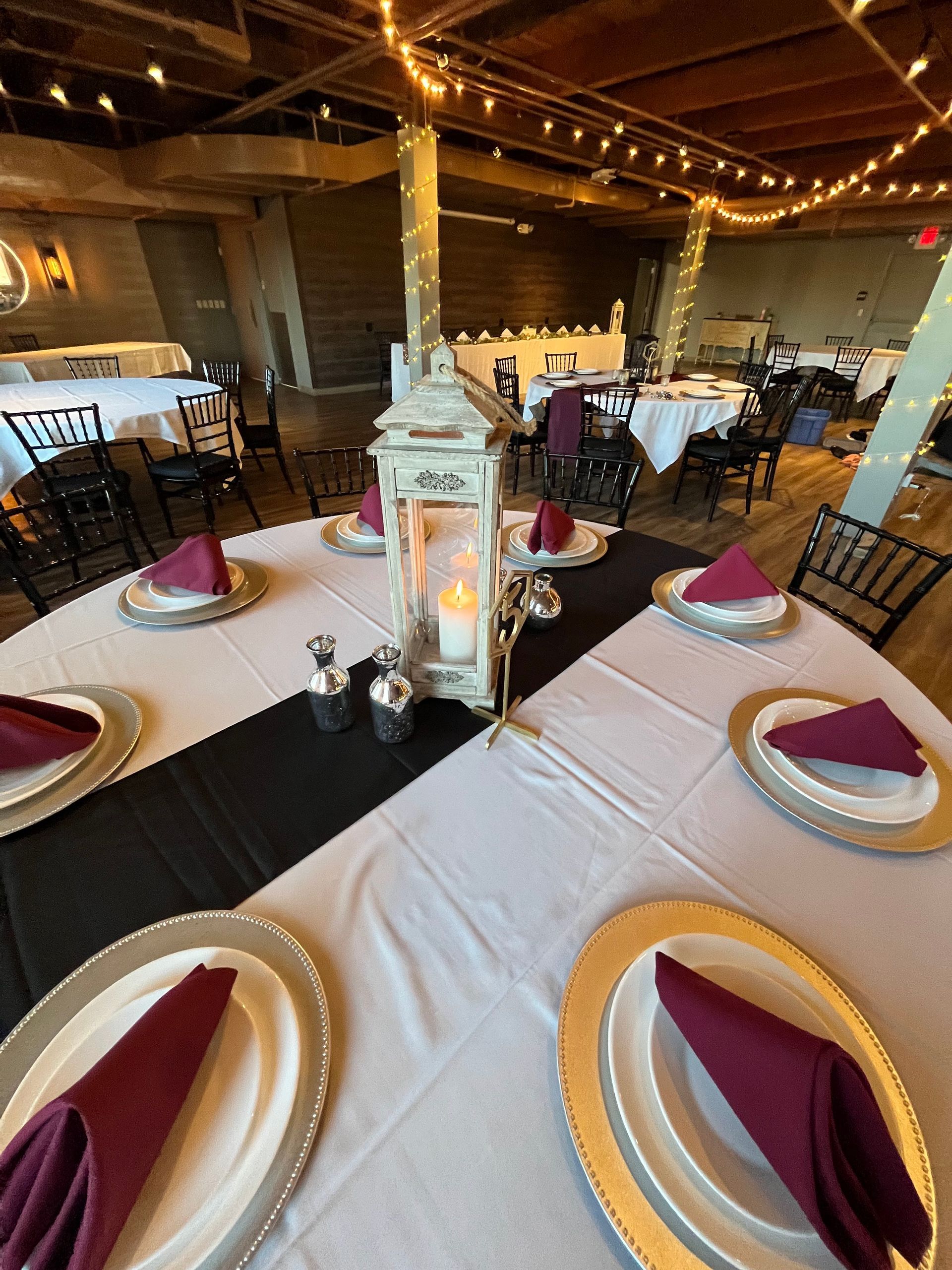 Banquet tables set with white cloths, black runners, gold chargers, and burgundy napkins in a rustic venue