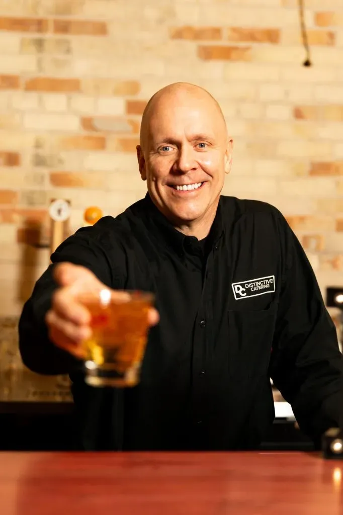 Smiling bartender in black shirt offering a drink behind a bar with a brick wall background