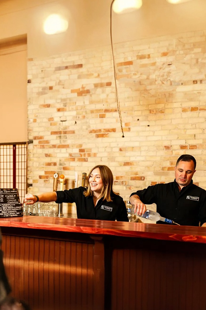 Two people seated behind a wooden bar counter in a warmly lit room with a tiled wall backdrop