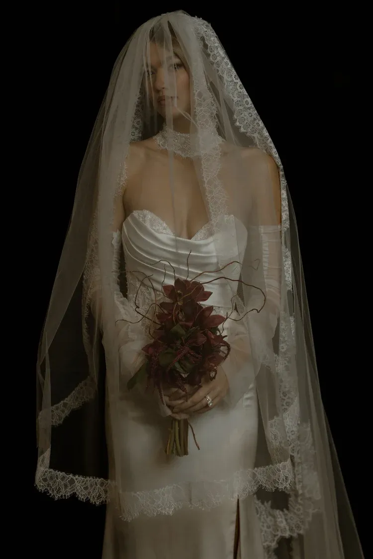 Bride in a white veil holding a bouquet of red flowers against a dark background