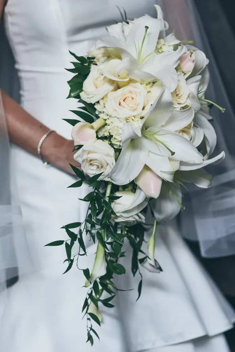 Bridal bouquet of white roses and lilies with trailing greenery, held against a white wedding dress