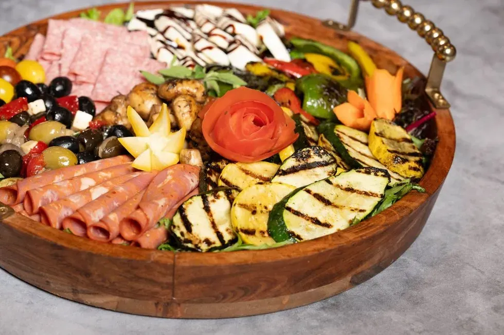 Wooden bowl of assorted fresh fruit, cheese, meats, and grilled vegetables on a gray surface