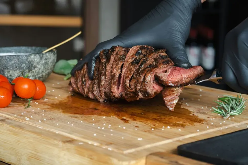 Gloved hand slicing a grilled steak on a wooden cutting board with tomatoes and rosemary nearby