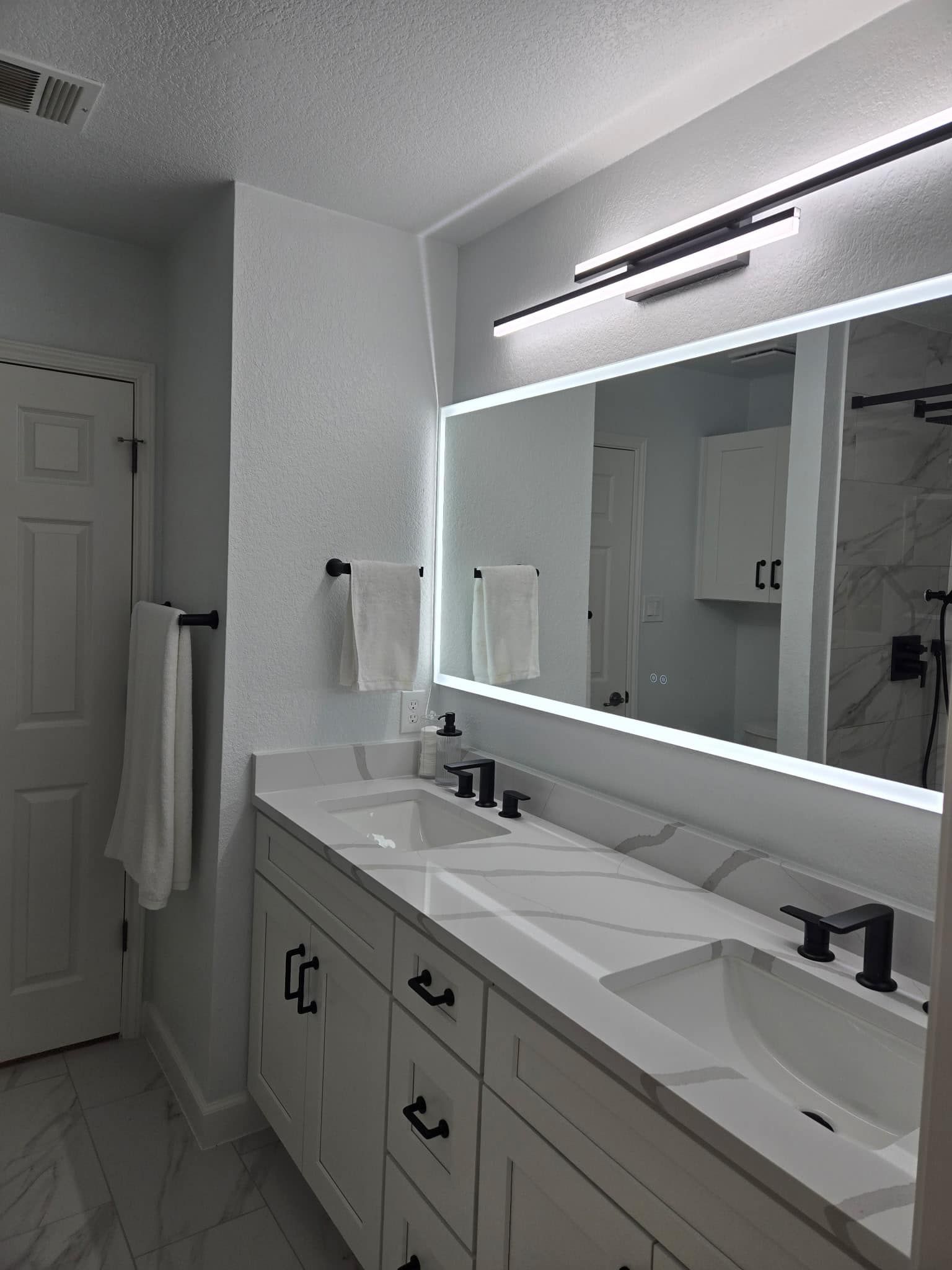 Modern white bathroom with double vanity, large illuminated mirror, and towel by the door