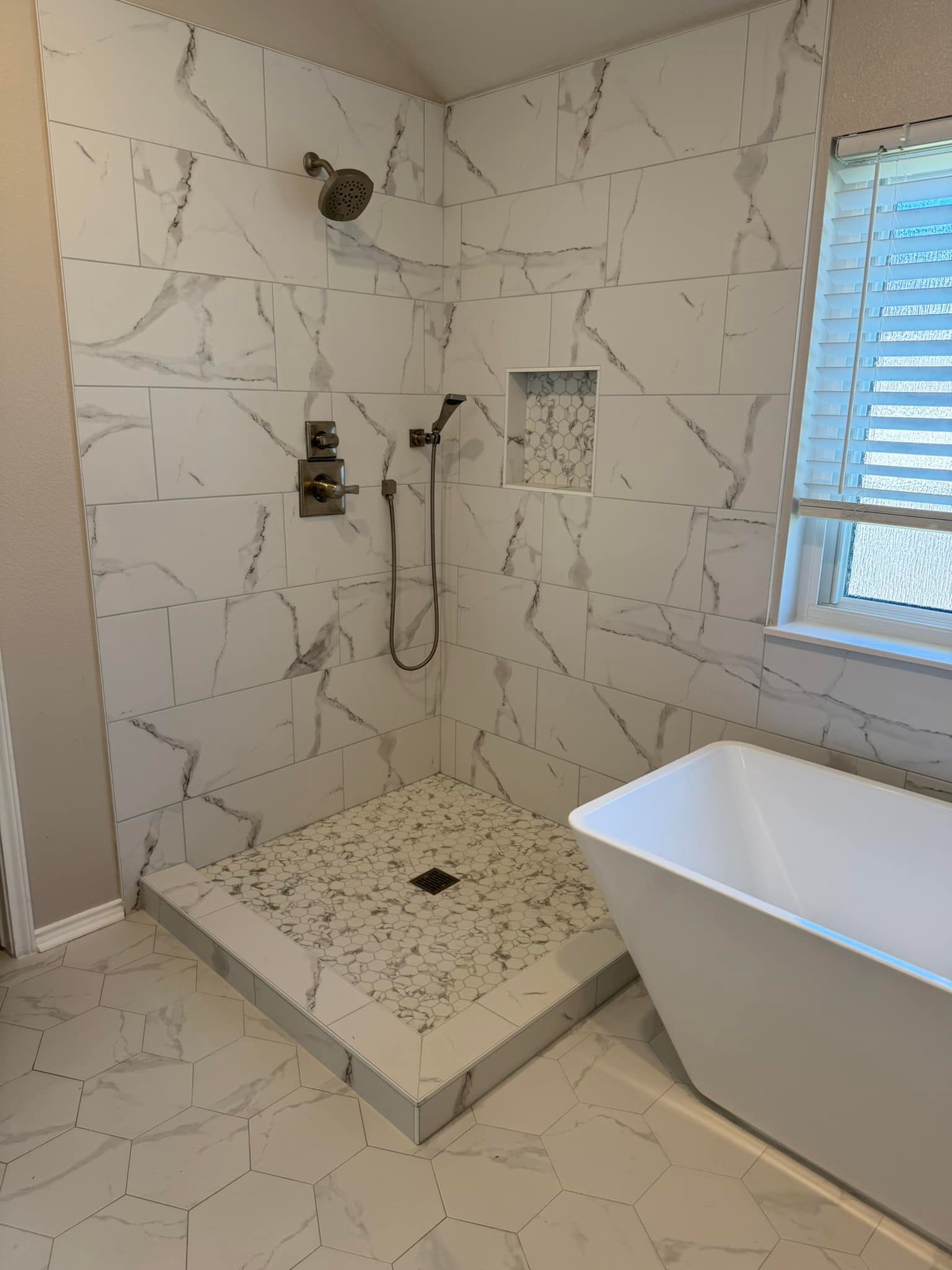 Modern bathroom shower with white marble tile, bench, handheld and fixed showerheads, beside a bathtub.