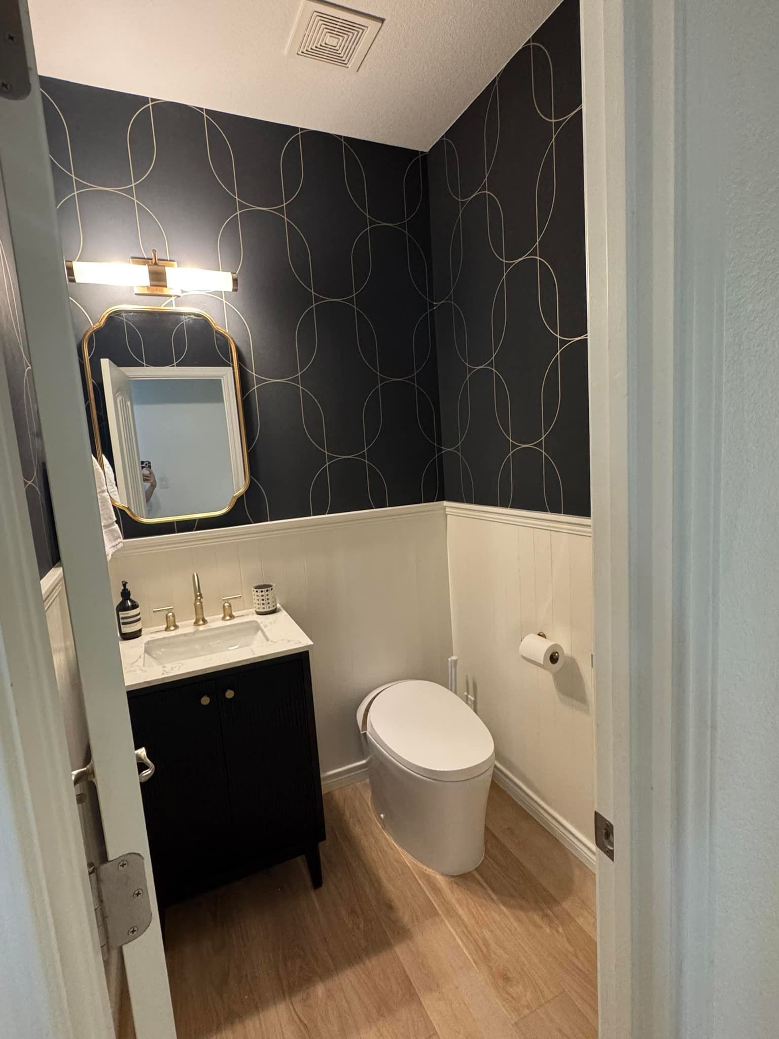 Small bathroom with dark patterned walls, white wainscoting, sink, mirror, toilet, and wood-look floor.