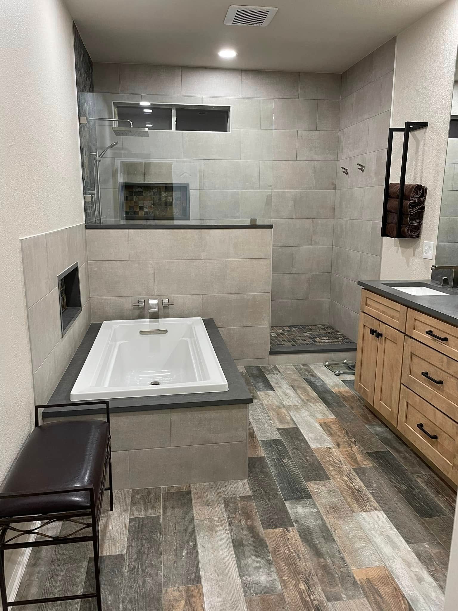 Modern bathroom with walk-in shower, white tub, wood vanity, and gray tiled walls and floor