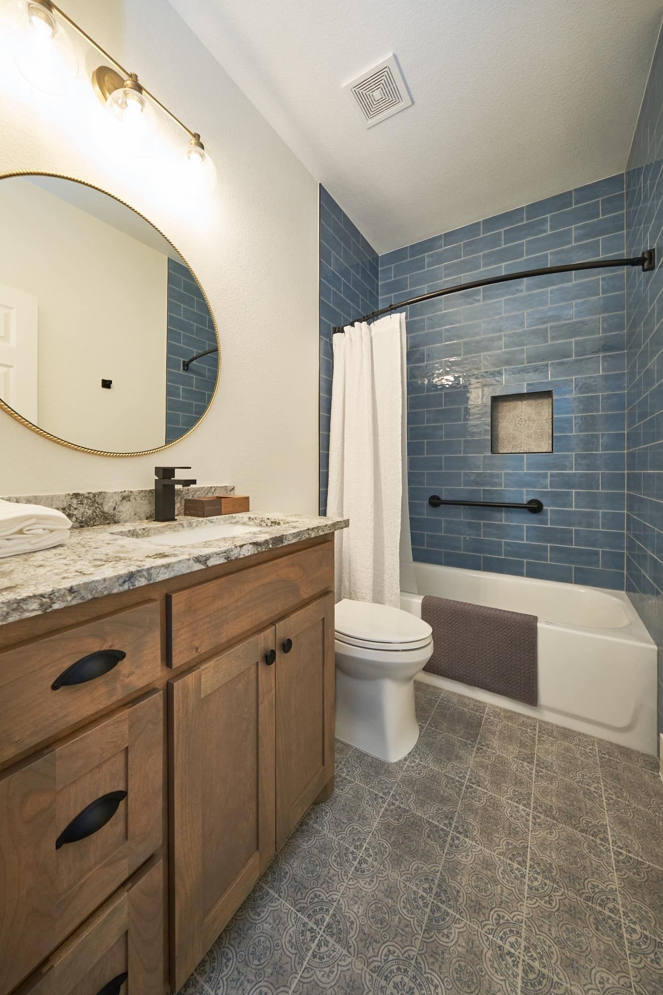 Modern bathroom with wood vanity, round mirror, blue tiled shower, and toilet
