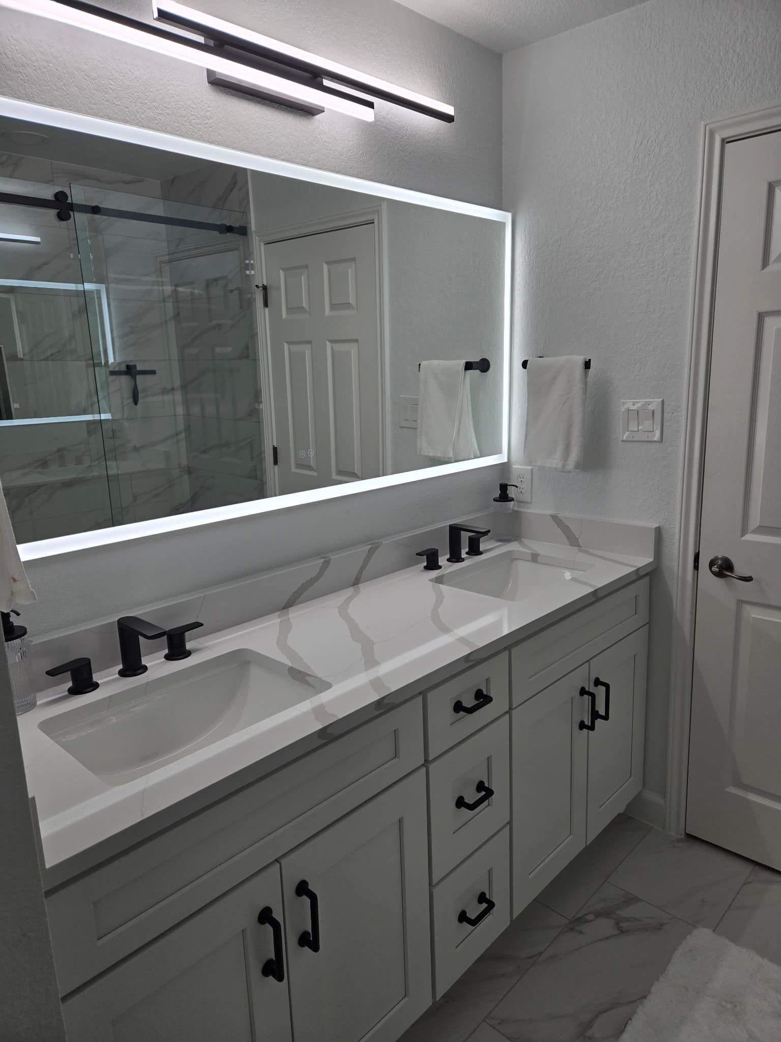 Modern bathroom vanity with double sinks, large lit mirror, white cabinets, and marble-style floor.