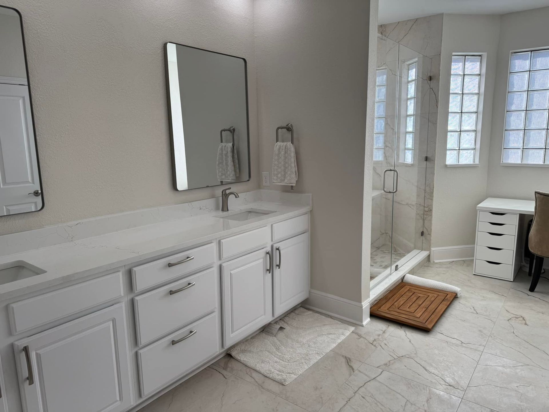Bright bathroom with white double vanity, mirror, tiled floor, and shower area by a windowed doorway