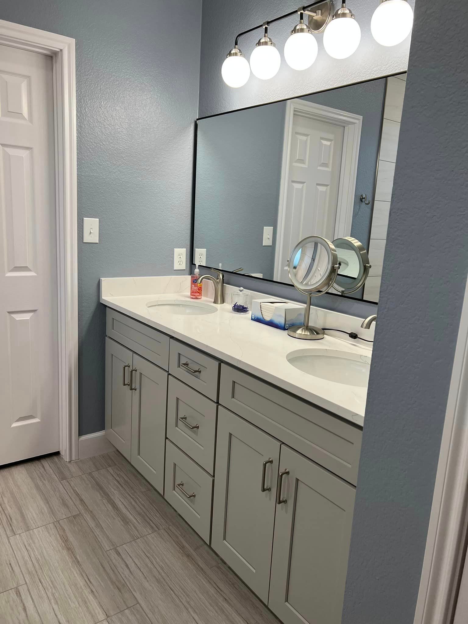 Bathroom vanity with gray cabinets, white countertop, large mirror, and bright overhead lights.