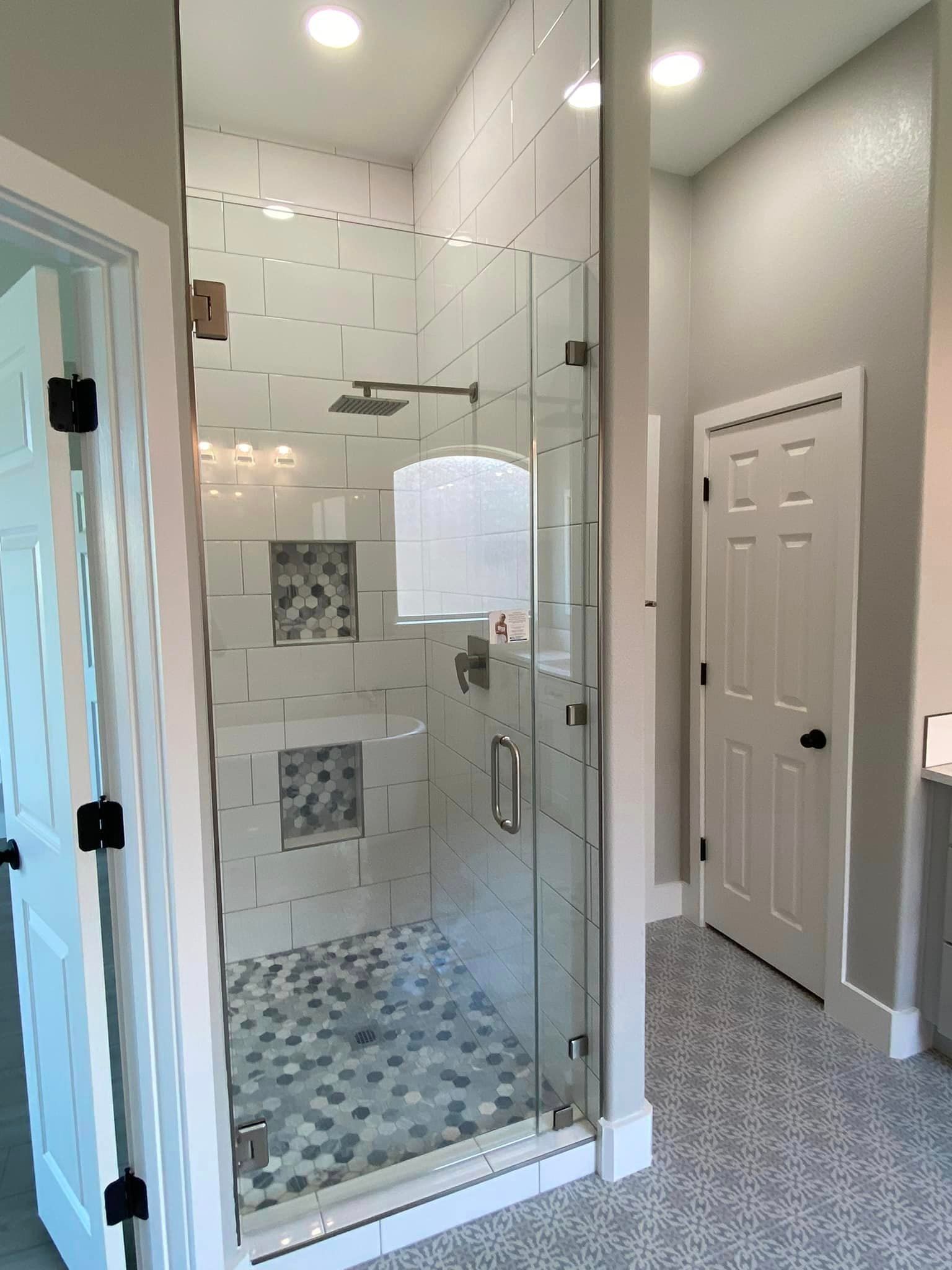 Glass walk-in shower in a bright bathroom with white tile and mosaic floor, adjacent to a carpeted hallway.