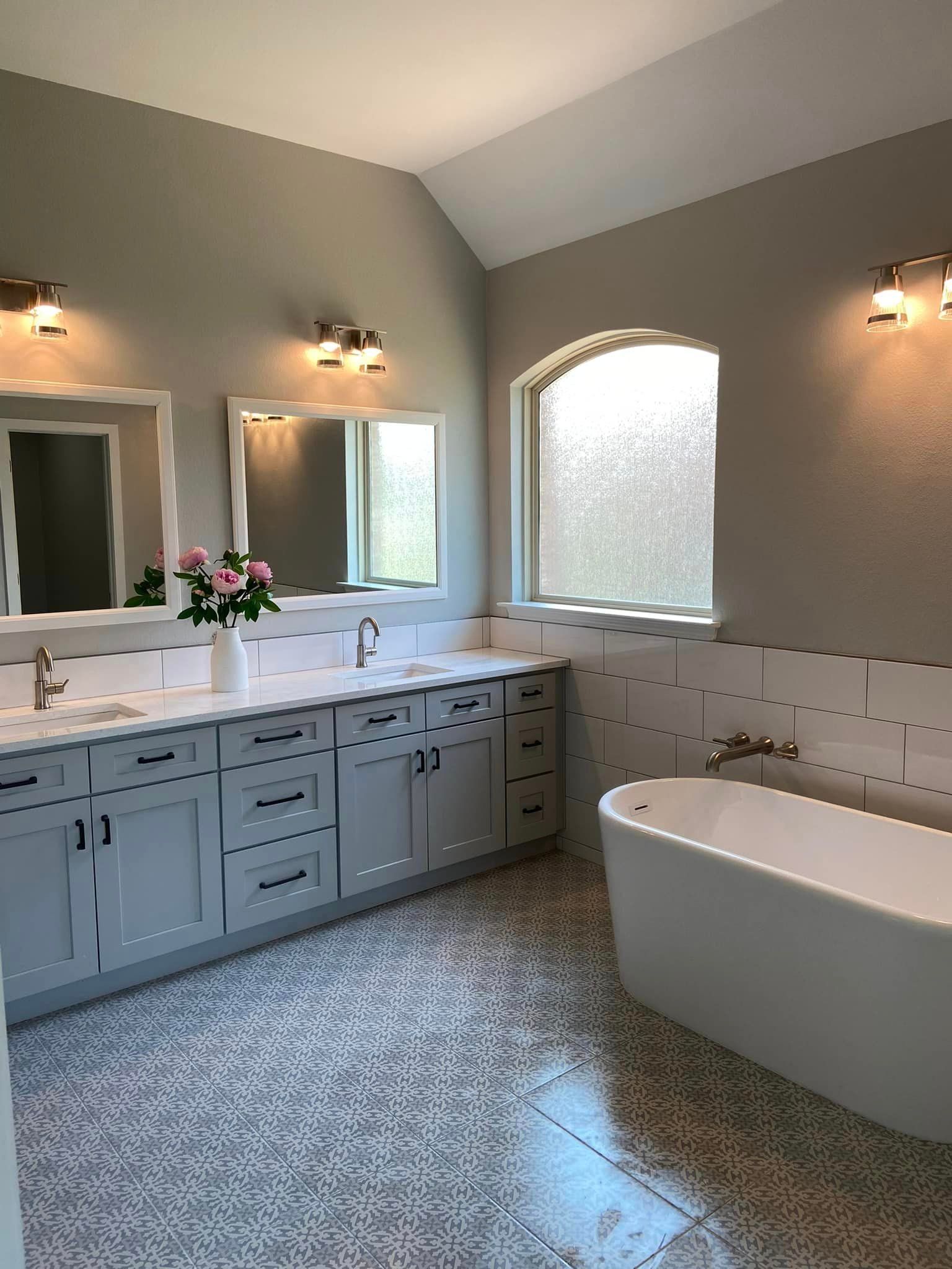 Bright bathroom with gray vanity, freestanding tub, double mirrors, and arched frosted window.