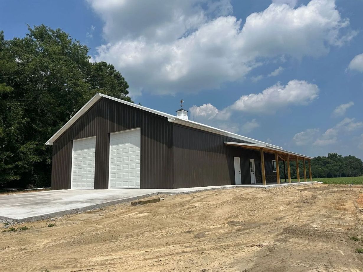 Large gray metal barn with open side bays on a dirt lot under a cloudy blue sky.