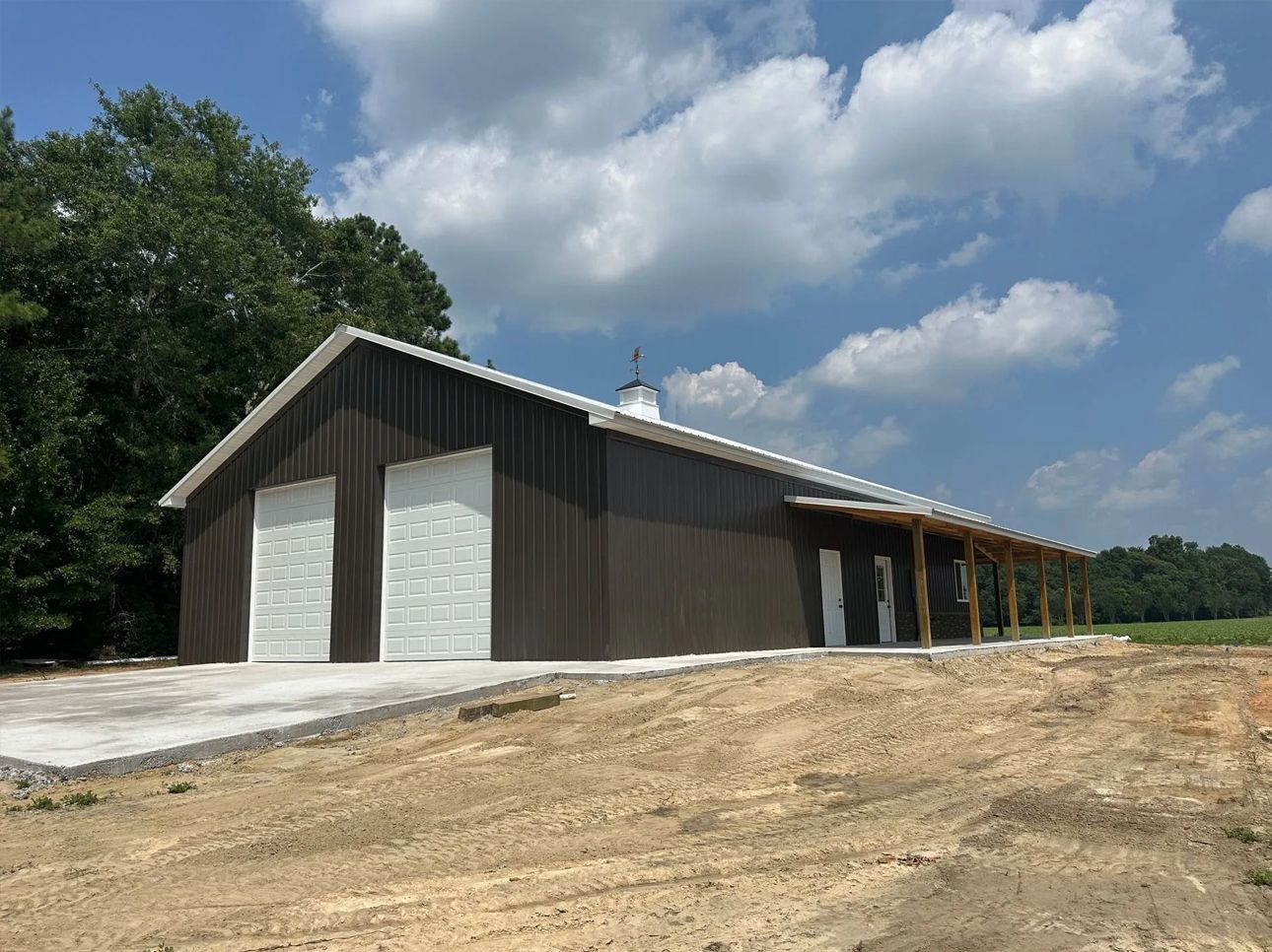 Large gray metal barn with open side bays on a dirt lot under a cloudy blue sky.