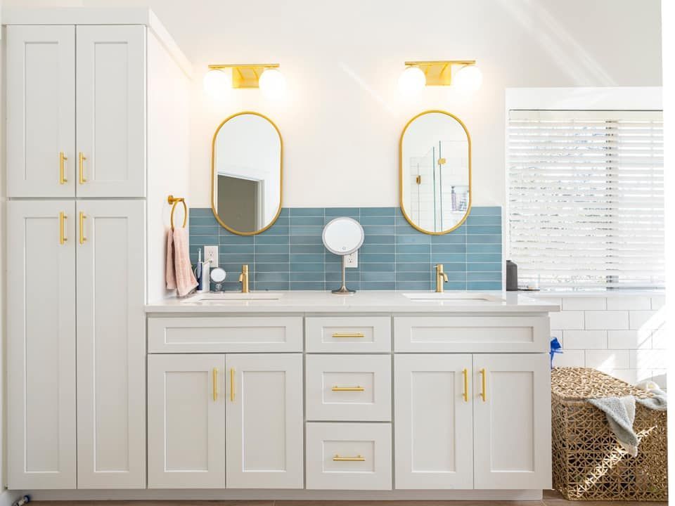 Bright white bathroom vanity with gold accents, blue tile backsplash, and two oval mirrors by a window.