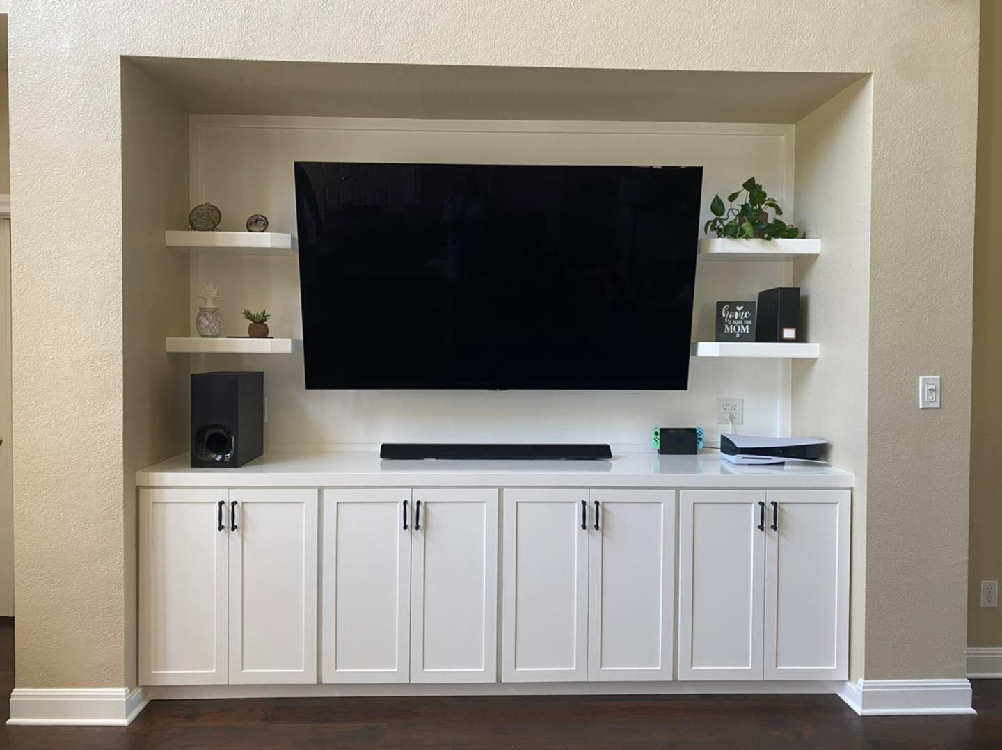 Built-in living room wall unit with large TV, white cabinets, and shelving in a beige alcove