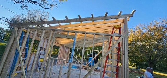 Partially built wooden house frame with scaffolding under a blue sky and autumn trees
