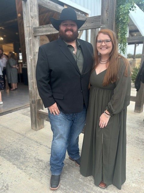 Two people posing together outdoors; one wears a black cowboy hat and blazer, the other a green dress.