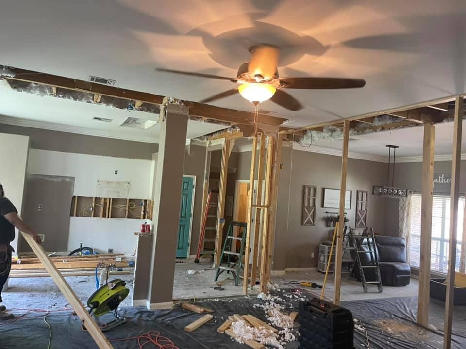 Living room under renovation with exposed studs, debris on floor, and ceiling fan overhead