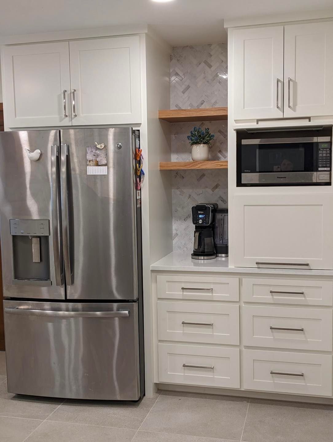 White kitchen with stainless steel fridge, built-in microwave, coffee maker, and white cabinets and drawers.
