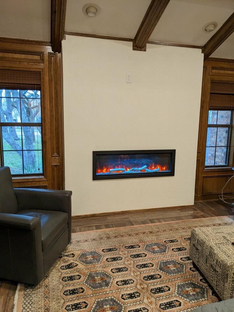 Cozy living room with a wall-mounted electric fireplace, windows, dark chair, and patterned rug