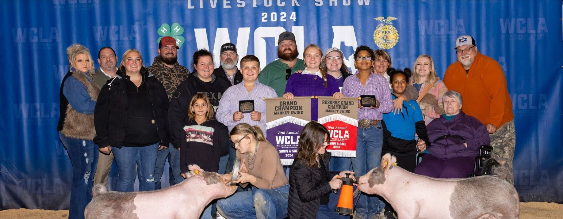 Group of people posing on a stage with award plaques and two pigs in front of a blue event backdrop