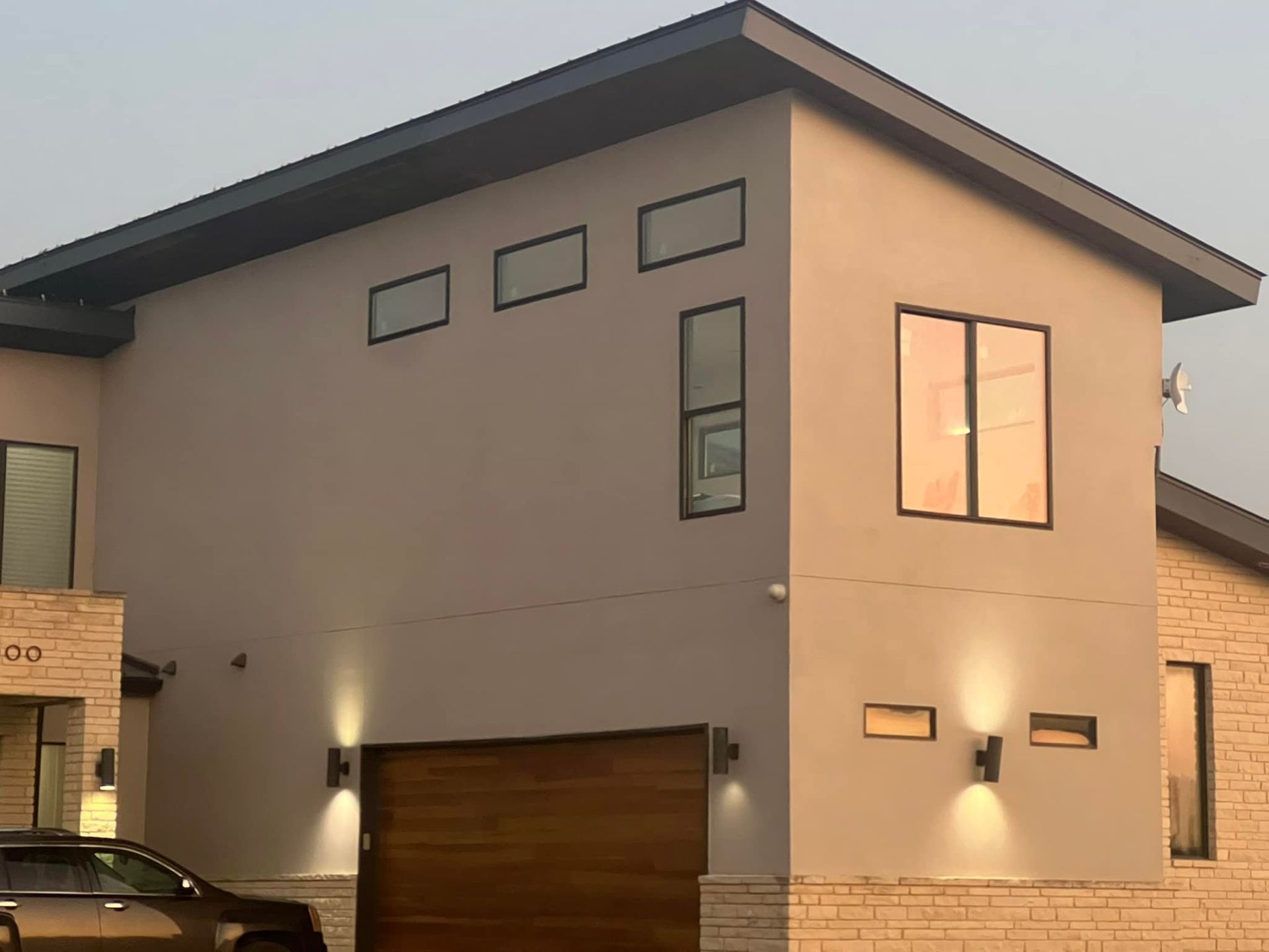 Modern gray two-story house with warm exterior lights and a wood garage door at dusk