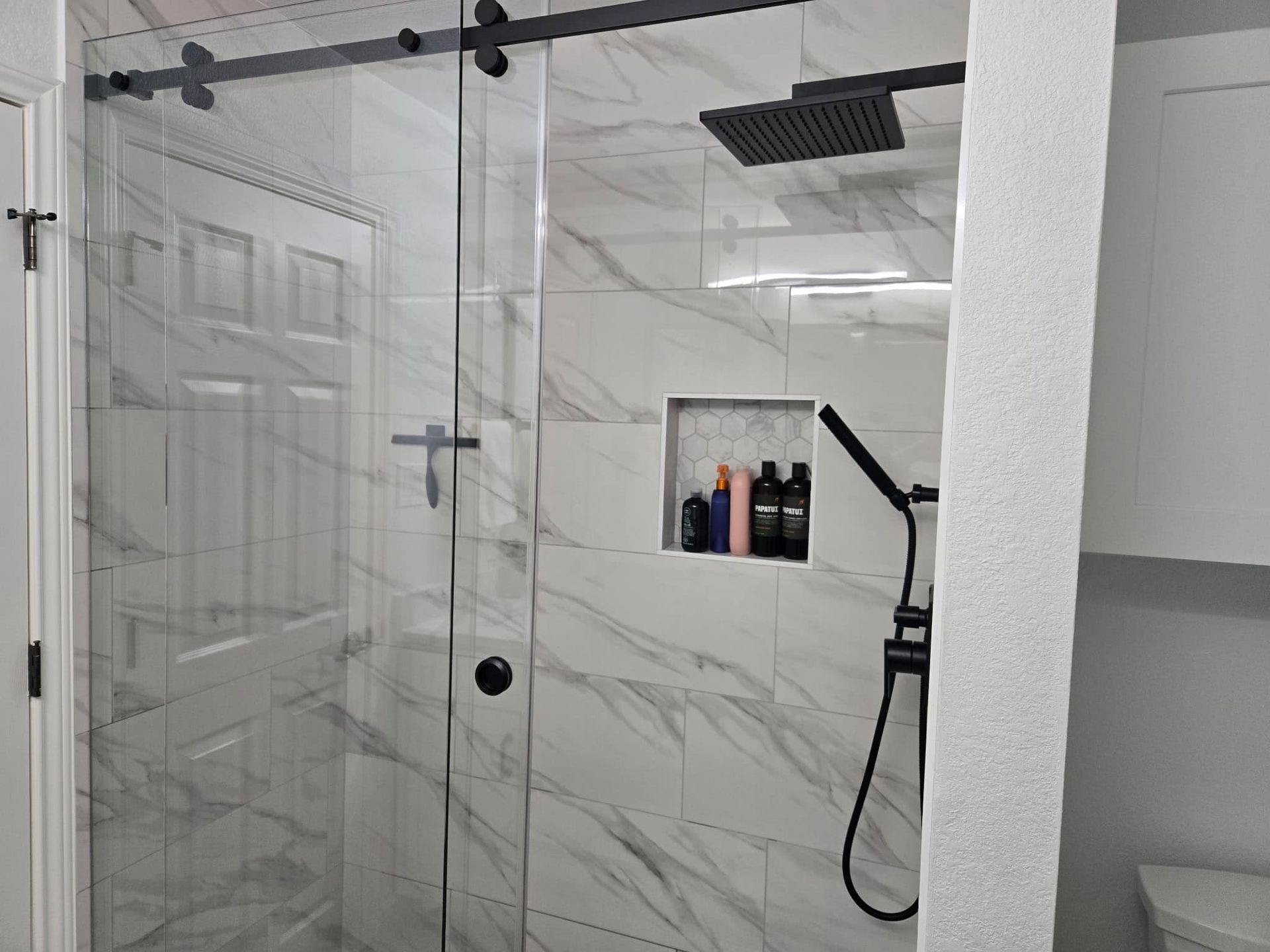 Modern shower with glass doors, marble-pattern tile, and a handheld showerhead with shelf of toiletries