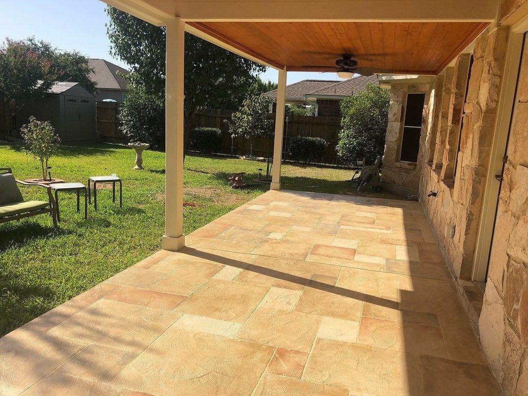 Covered stone patio beside a grassy backyard, with a wooden roof and stone wall in sunlight