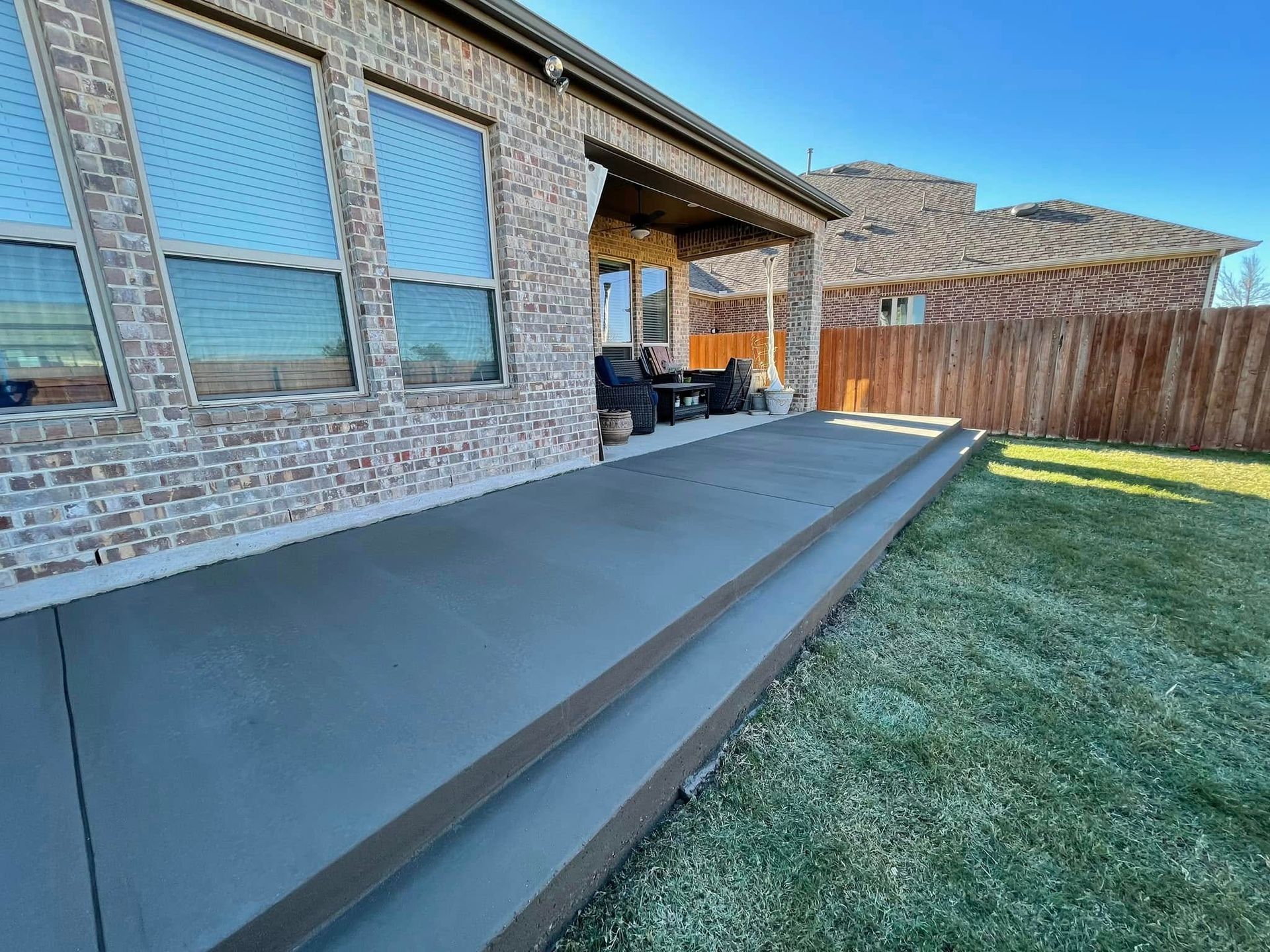 Backyard patio with a long concrete step beside a brick house and grass lawn, with a wooden fence.