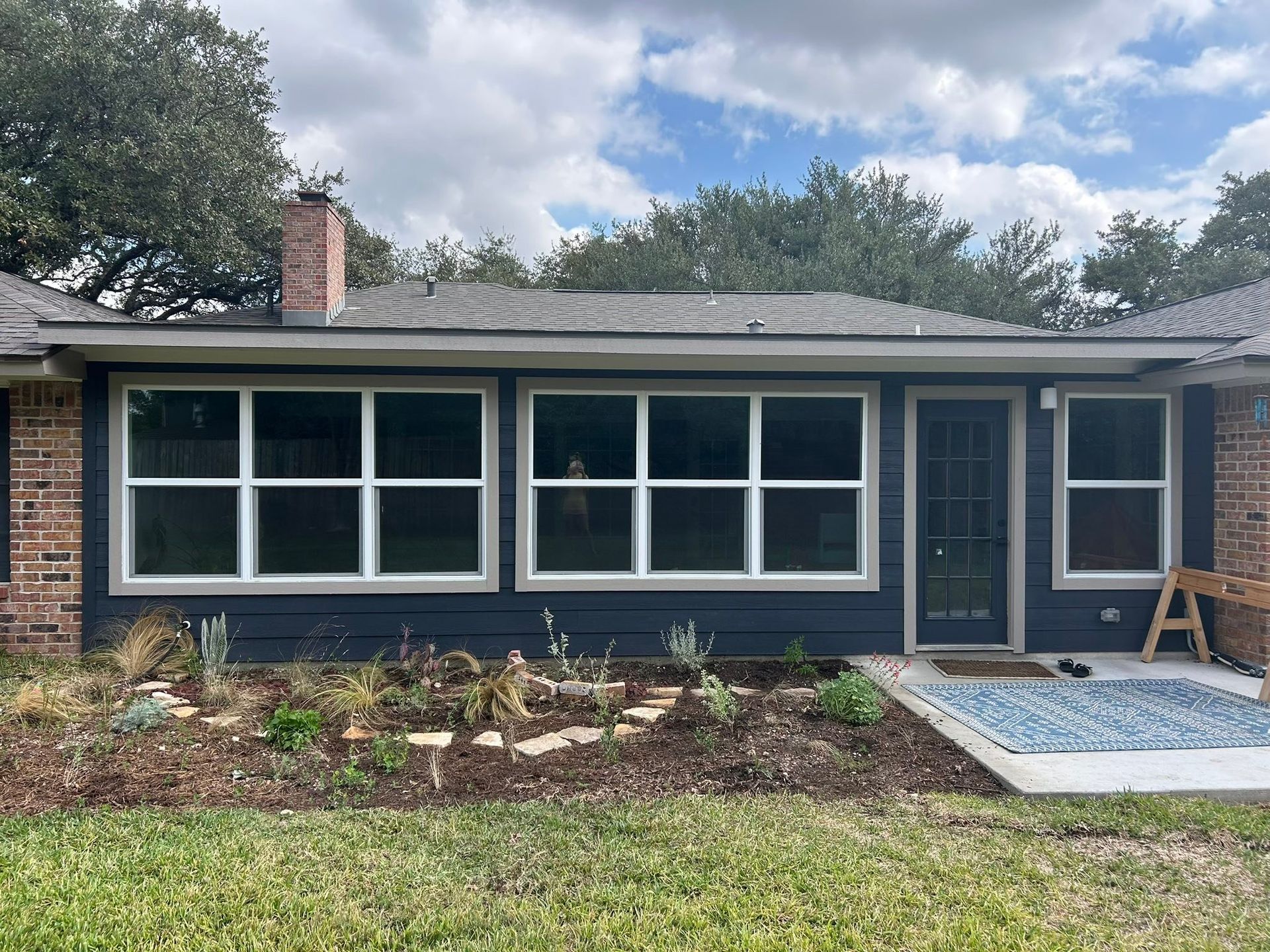 Single-story brick house with dark blue siding, large windows, front door, and garden beds under a cloudy sky