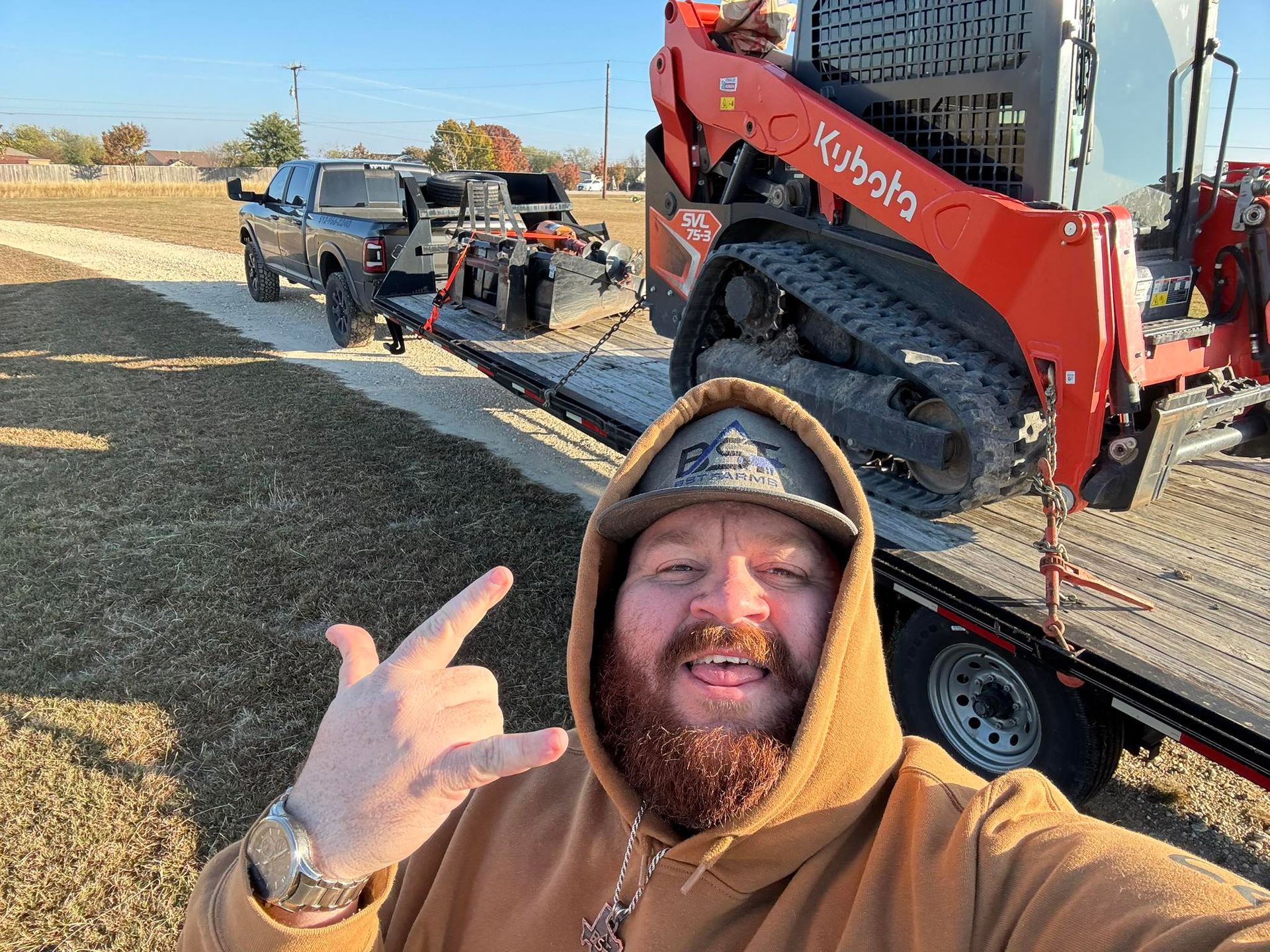 Smiling bearded man in brown hoodie posing beside a red Bobcat loader on a trailer outdoors