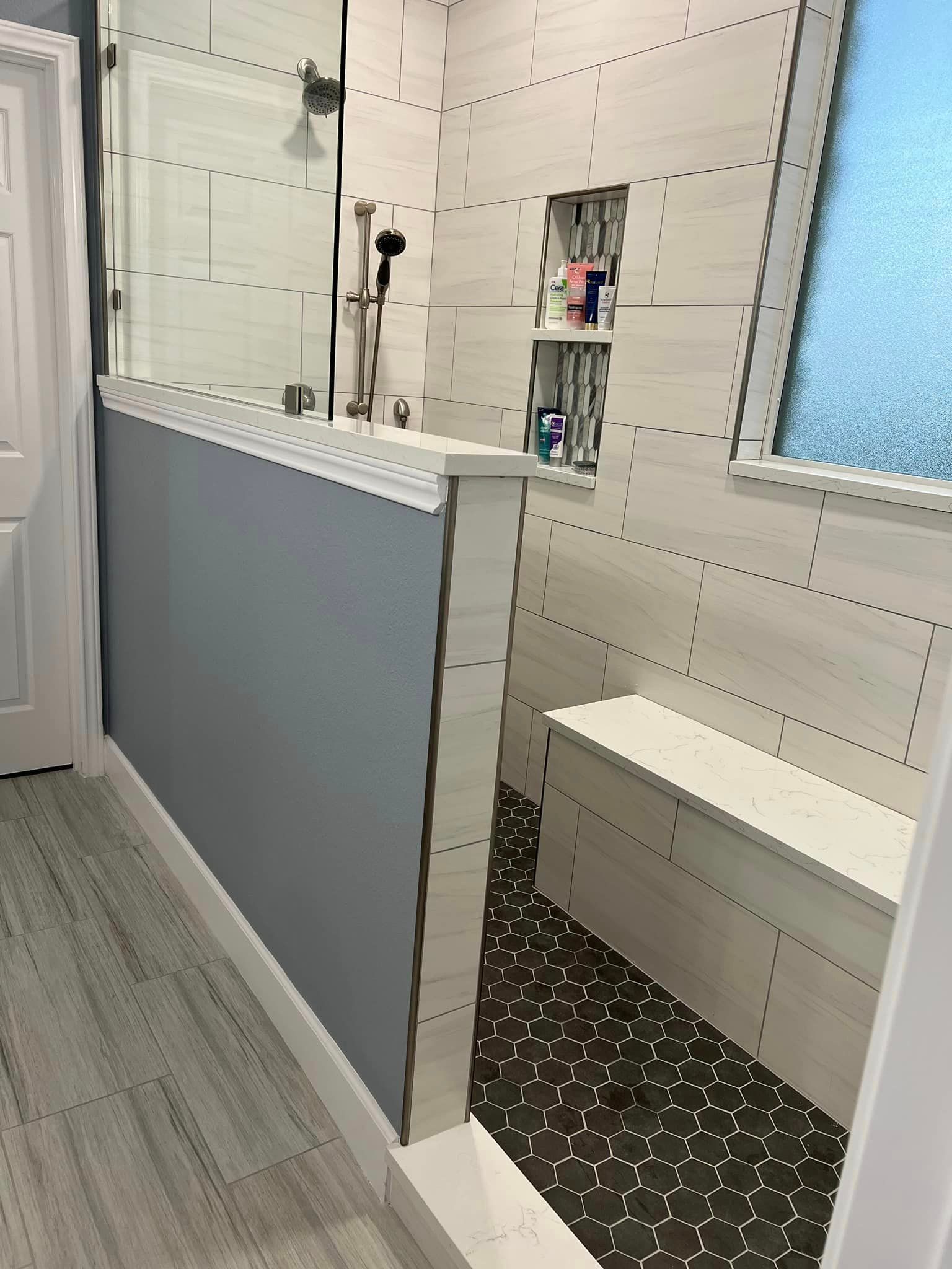 Small bathroom with shower, sink, toilet, and a white bench on patterned flooring.