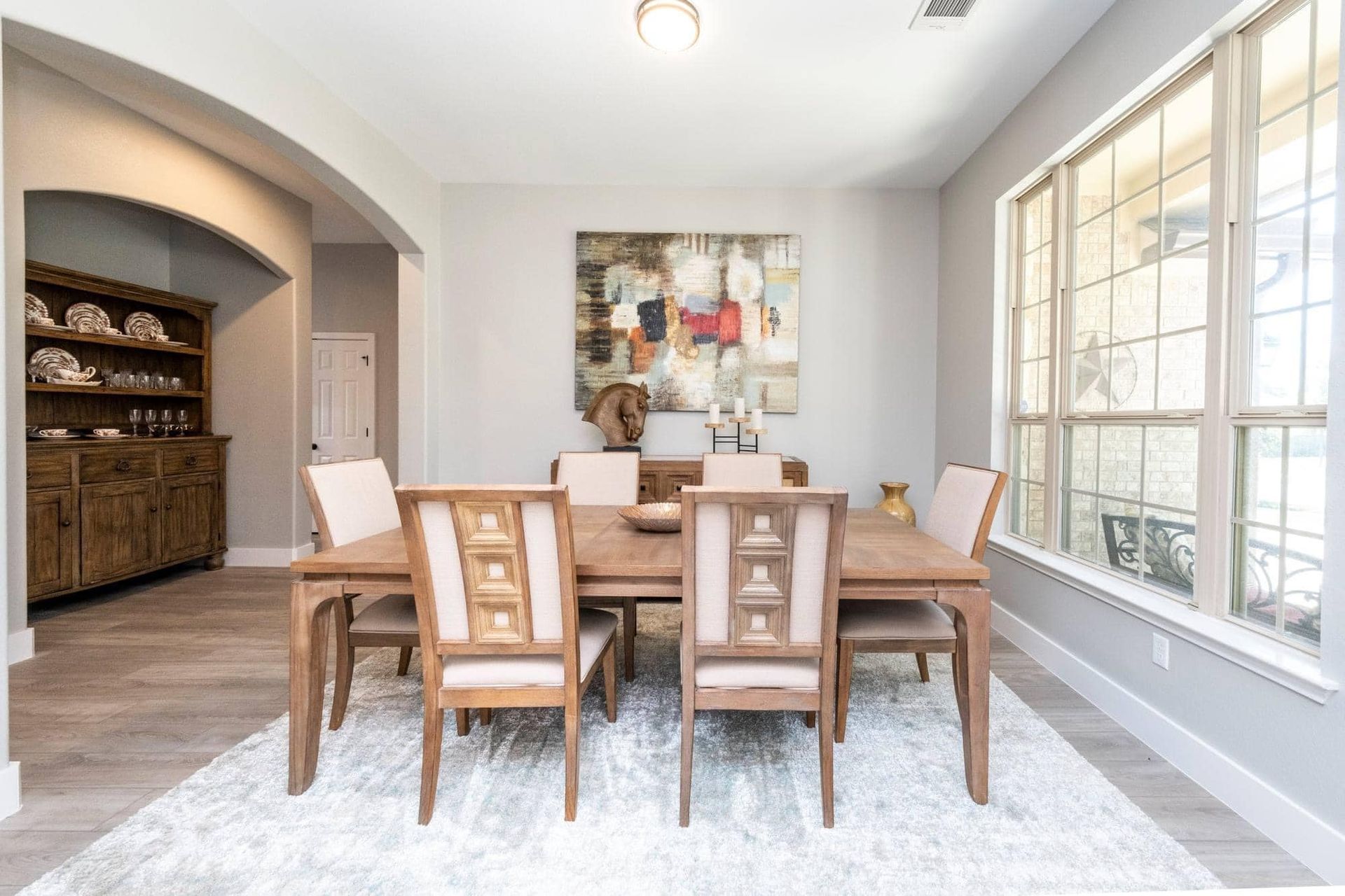 Bright dining room with wooden table and chairs, white rug, large windows, and wall art.