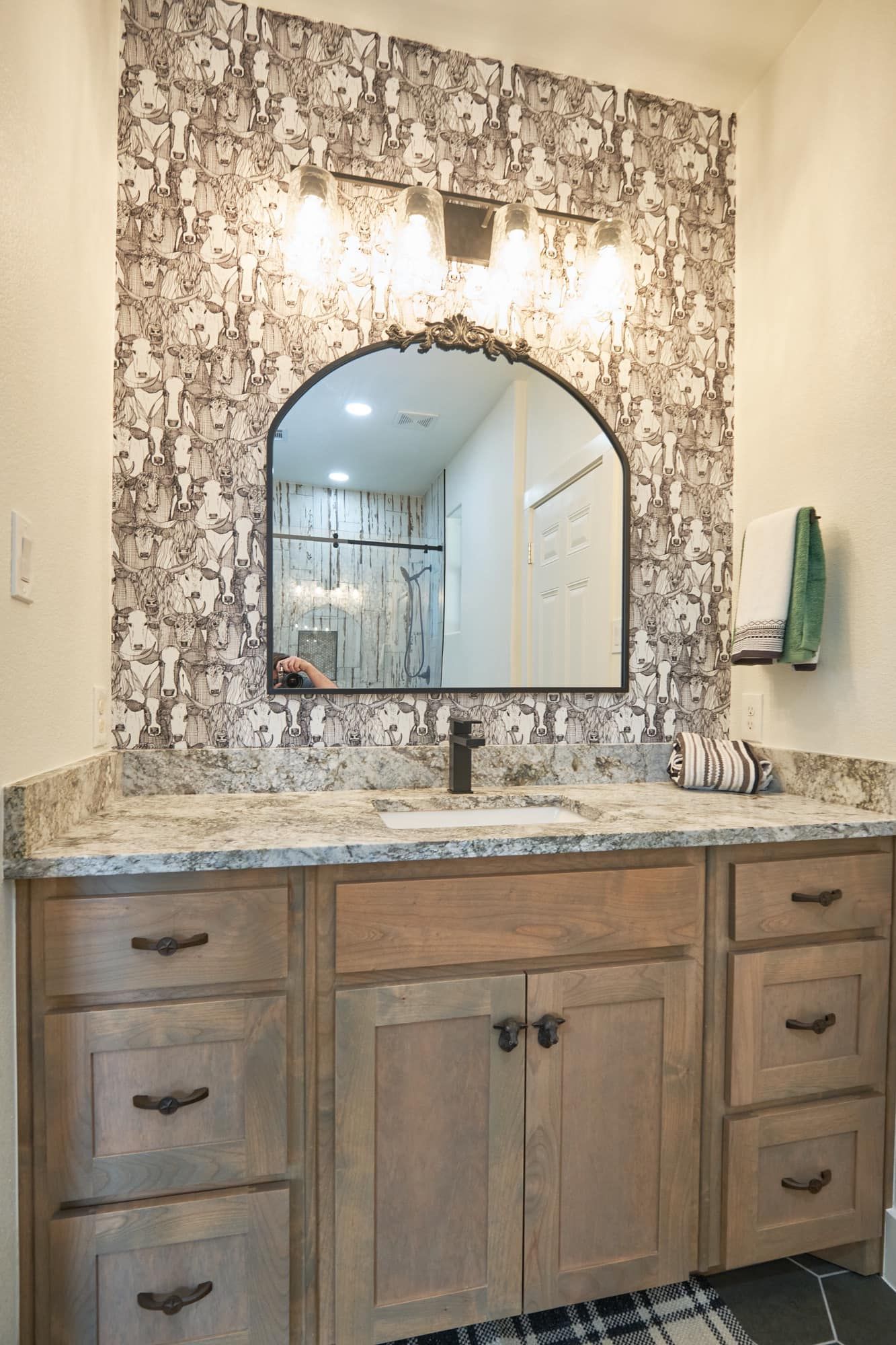 Bathroom vanity with granite countertop, arched mirror, and wall lights against patterned backsplash.