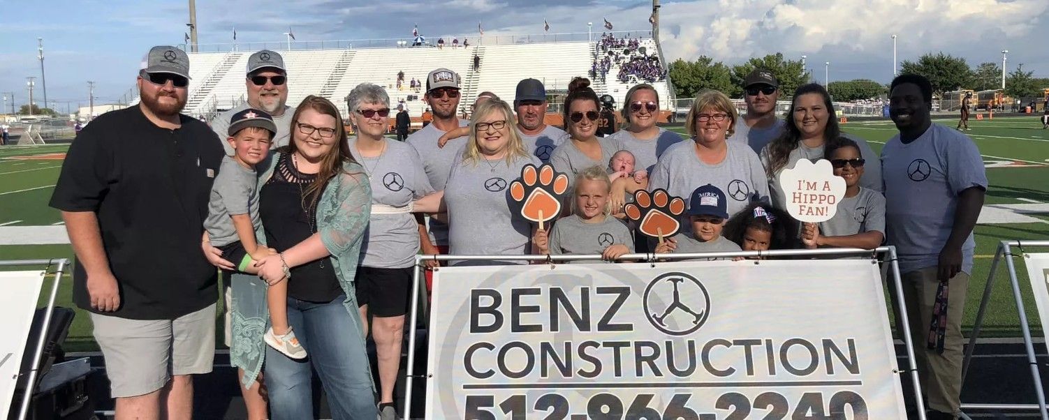 Group posing outdoors behind a Benz Construction sign, many holding orange cutouts.