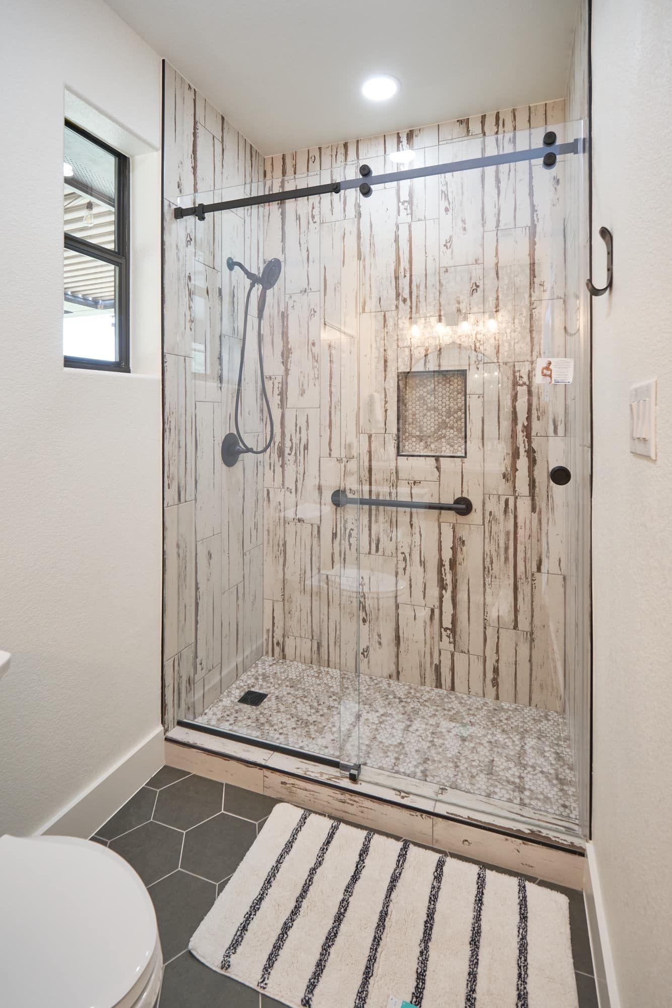 Modern bathroom shower with wood-look tile, black fixtures, and a striped bath mat