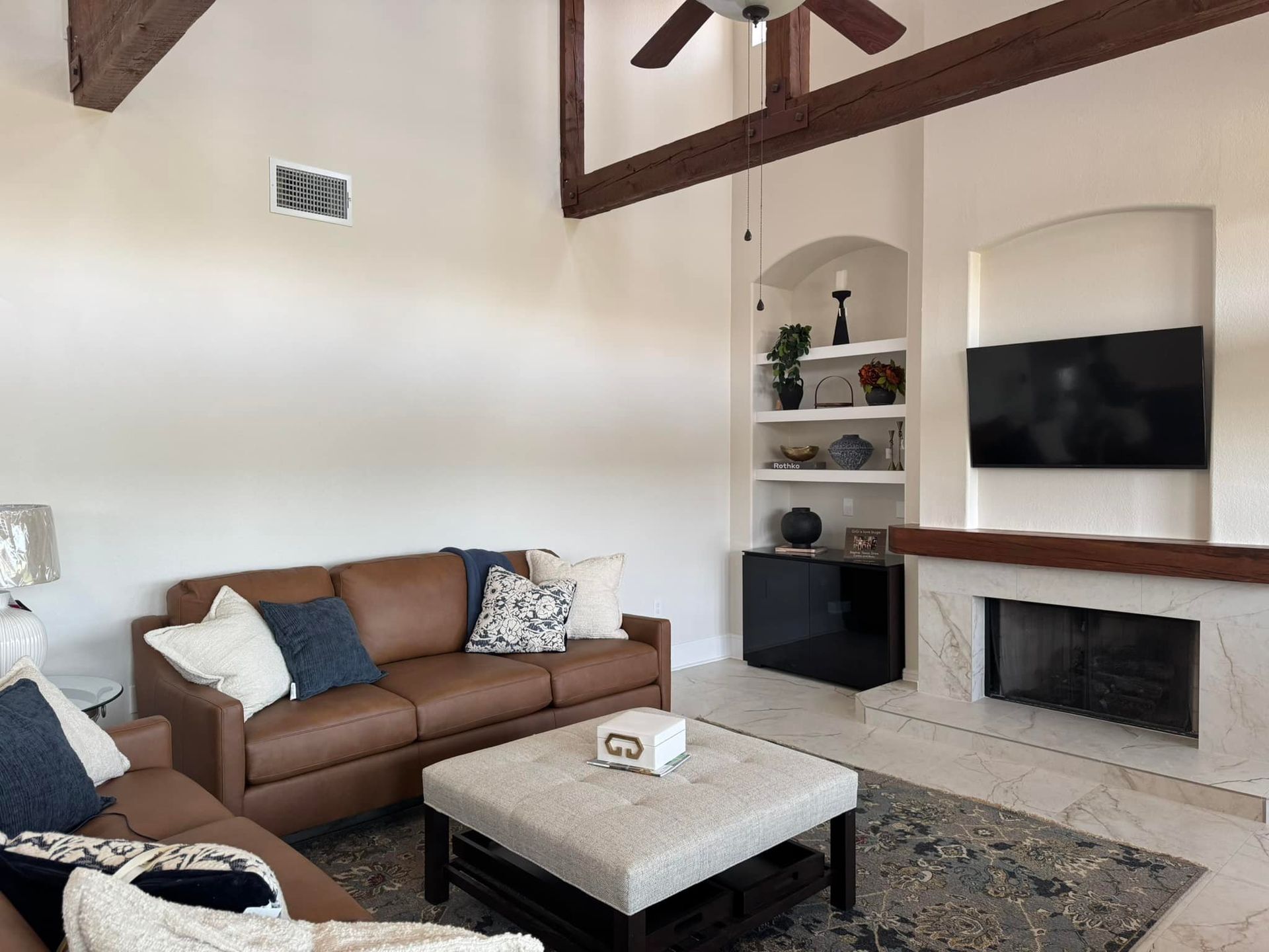 Modern living room with brown sofa, white fireplace, built-in shelves, and exposed wood beams.