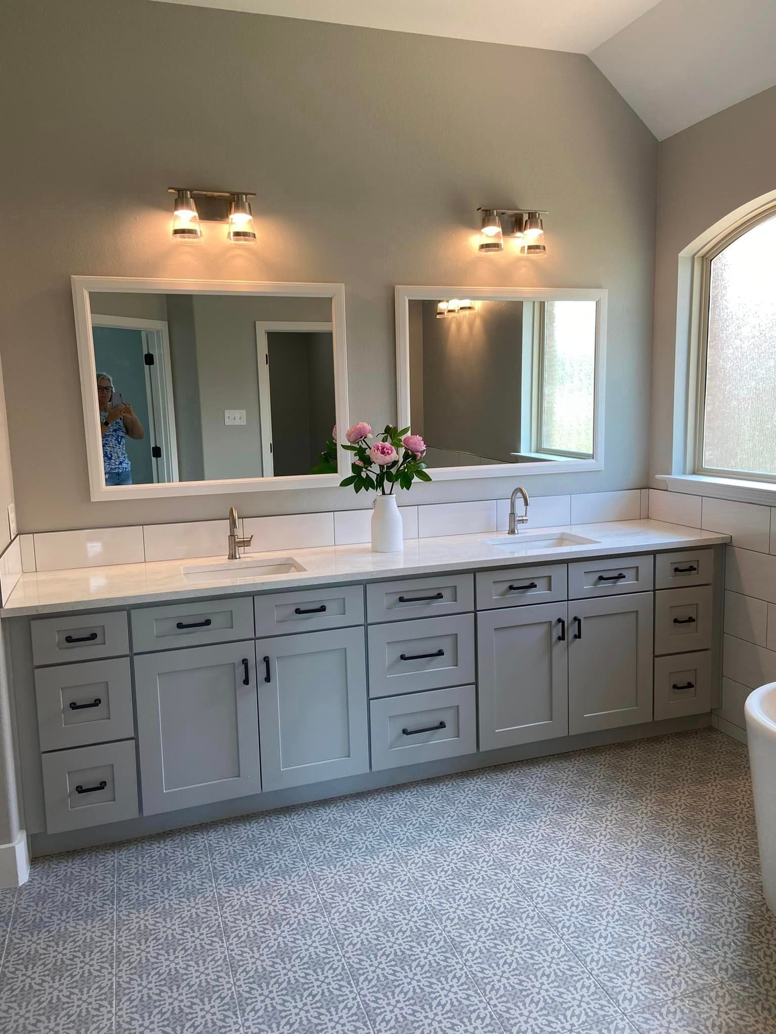 Gray bathroom vanity with double mirrors, wall sconces, white countertop, and a vase of pink flowers
